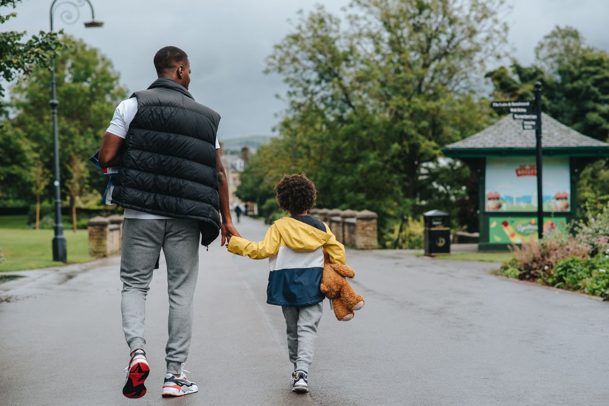 A guardian and a young child walk hand in hand along a tree-lined path