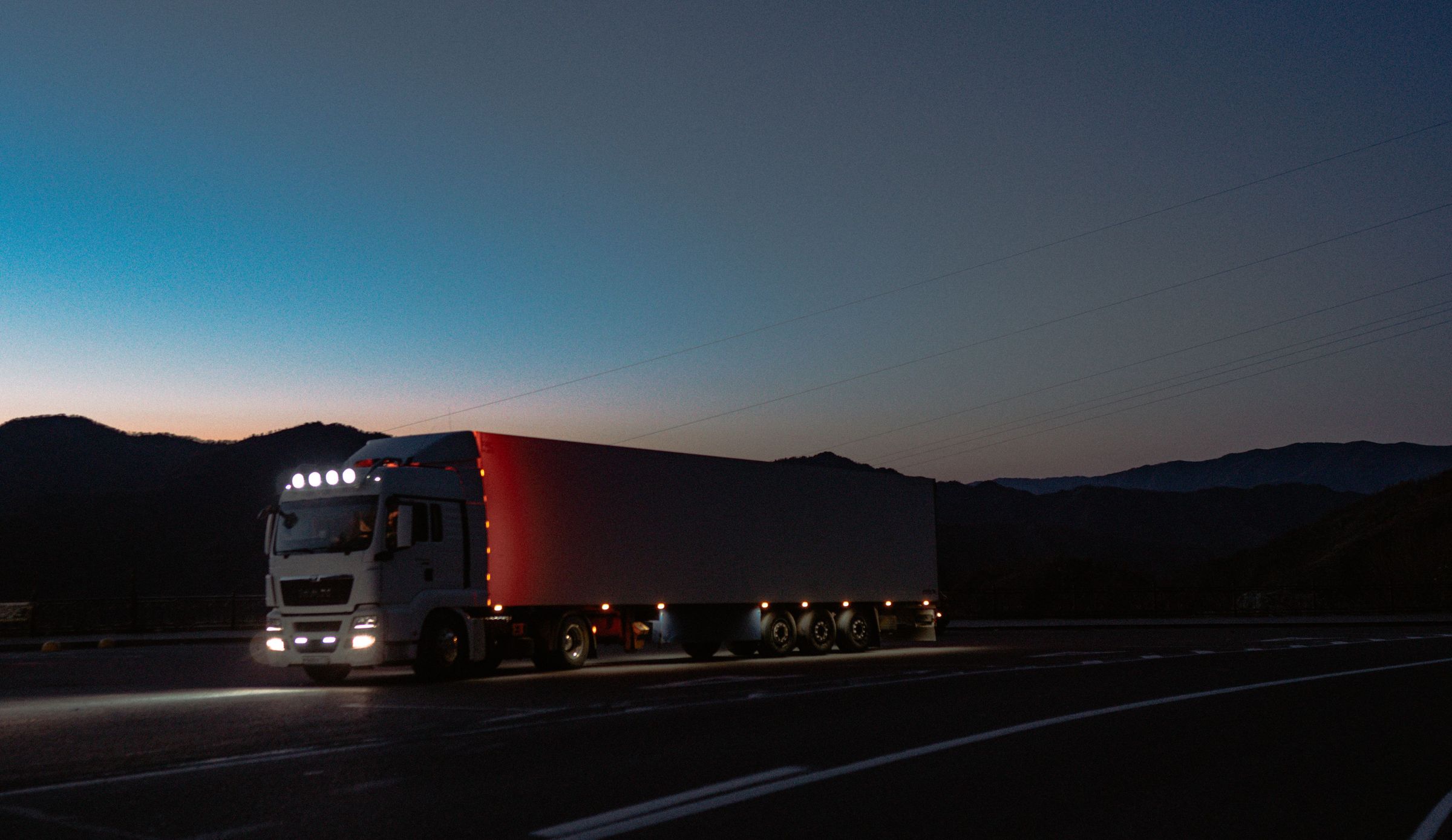A semi truck on a dark road at dusk, running lights on, mountains behind